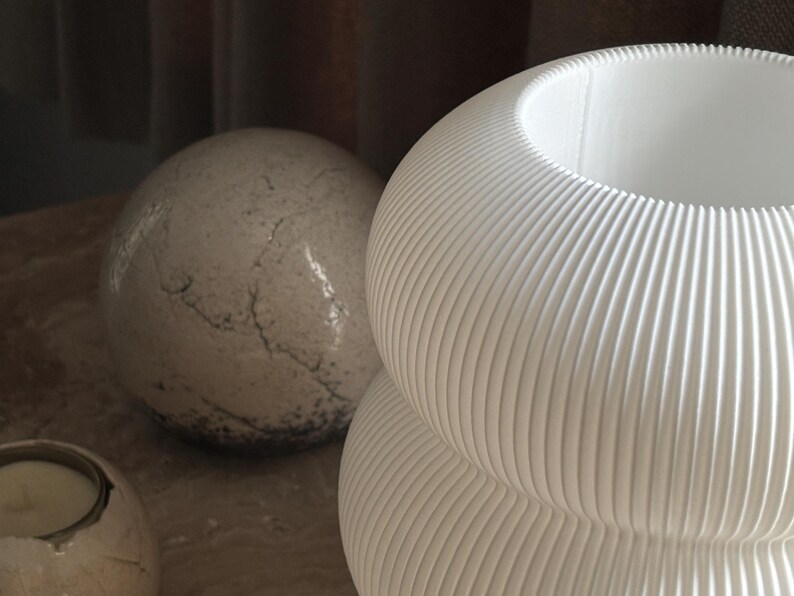 May include: A white vase with a rounded, stacked shape and a vertical ribbed texture. Beside it sits a spherical object with a mottled, stone-like surface featuring dark grey cracks and a small, circular candle holder with a metallic rim.