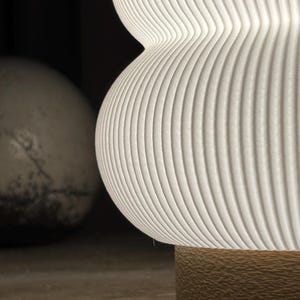 May include: A white, ribbed, hourglass-shaped lamp shade with a textured surface. The shade is illuminated from within, casting a soft glow. Below the shade is a cylindrical base with a rough, tan, granular texture, providing a contrast.