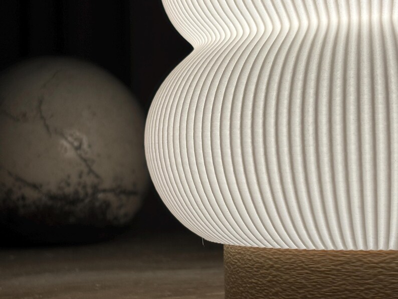 May include: A white, ribbed, hourglass-shaped lamp shade with a textured surface. The shade is illuminated from within, casting a soft glow. Below the shade is a cylindrical base with a rough, tan, granular texture, providing a contrast.