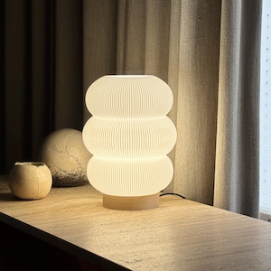 May include: A white, ribbed, three-tiered cylindrical table lamp with a glowing light effect, mounted on a circular light-colored wooden base. The lamp features a distinct stacked, rounded shape with vertical texture lines throughout the surface.