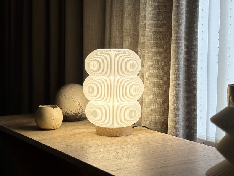 May include: A white, ribbed, three-tiered cylindrical table lamp with a glowing light effect, mounted on a circular light-colored wooden base. The lamp features a distinct stacked, rounded shape with vertical texture lines throughout the surface.