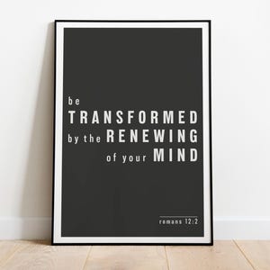 May include: A framed poster with a black background and white text. The text reads "be TRANSFORMED by the RENEWING of your MIND" with "Romans 12:2" at the bottom. The poster is displayed on a wooden floor.