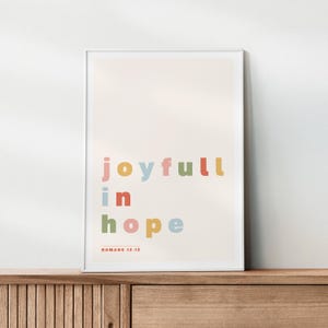 May include: A framed print with the words "joyfull in hope" in colorful block letters. The text "Romans 12:12" is printed below the words. The background is a light beige color.