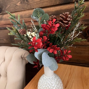 Puede incluir: Un jarrón de cerámica blanco con una superficie texturizada contiene una disposición festiva de flores rojas con purpurina, hojas de eucalipto verdes y piñas marrones.