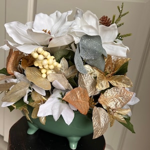 Puede incluir: Un arreglo floral blanco y dorado en una maceta de cerámica verde con tres patas. El arreglo incluye flores blancas de magnolia, hojas doradas y marrones y bayas blancas.