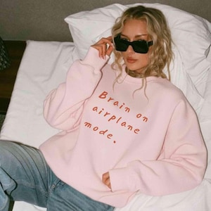 K&ouml;nnte beinhalten: Ein hellrosa Sweatshirt mit dem Text "Brain on airplane mode" in Rot. Das Sweatshirt wird mit einer blauen Jeans kombiniert. Die Person auf dem Bild tr&auml;gt eine schwarze Sonnenbrille.