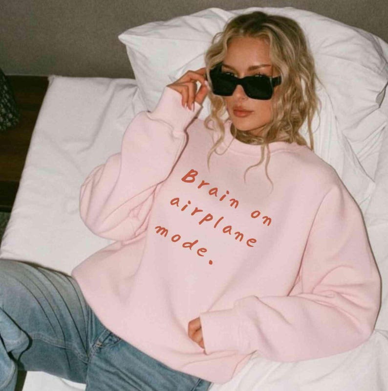 K&ouml;nnte beinhalten: Ein hellrosa Sweatshirt mit dem Text "Brain on airplane mode" in Rot. Das Sweatshirt wird mit einer blauen Jeans kombiniert. Die Person auf dem Bild tr&auml;gt eine schwarze Sonnenbrille.