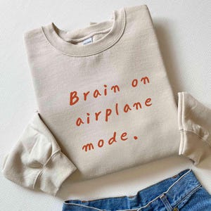 K&ouml;nnte beinhalten: Beiges Sweatshirt mit dem Text "Brain on airplane mode" in orangefarbenen Buchstaben. Das Sweatshirt ist gefaltet, die &Auml;rmel sind sichtbar. Blaue Jeans sind am unteren Bildrand zu sehen.