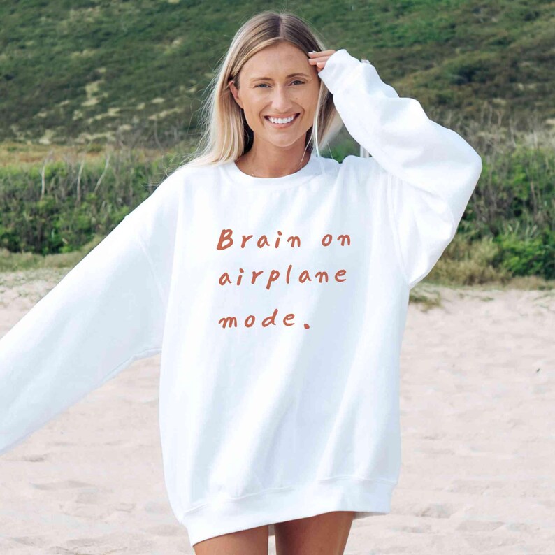 K&ouml;nnte beinhalten: Wei&szlig;es Sweatshirt mit dem Text "Brain on airplane mode" in Rotbraun. Das Sweatshirt mit langen &Auml;rmeln und Rundhalsausschnitt wird im Freien getragen. Das Bild wird an einem Sandstrand aufgenommen.