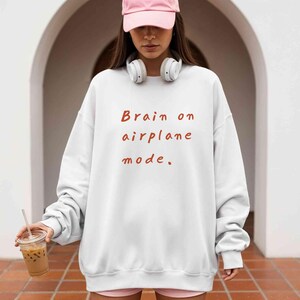 K&ouml;nnte beinhalten: Wei&szlig;es Sweatshirt mit dem Text "Brain on airplane mode" in Rotbraun. Das Sweatshirt hat lange &Auml;rmel und einen Rundhalsausschnitt. Das Bild zeigt eine Person, die das Sweatshirt, eine rosa Kappe und wei&szlig;e Kopfh&ouml;rer tr&auml;gt.