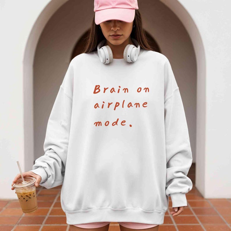 K&ouml;nnte beinhalten: Wei&szlig;es Sweatshirt mit dem Text "Brain on airplane mode" in Rotbraun. Das Sweatshirt hat lange &Auml;rmel und einen Rundhalsausschnitt. Das Bild zeigt eine Person, die das Sweatshirt, eine rosa Kappe und wei&szlig;e Kopfh&ouml;rer tr&auml;gt.
