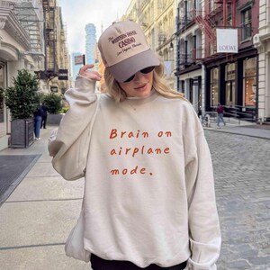 K&ouml;nnte beinhalten: Beiges Sweatshirt mit dem Text "Brain on airplane mode" in roten Buchstaben. Die Person tr&auml;gt eine beige und graue Baseballkappe mit dem Aufdruck "The Western House Casino Las Vegas Florida".