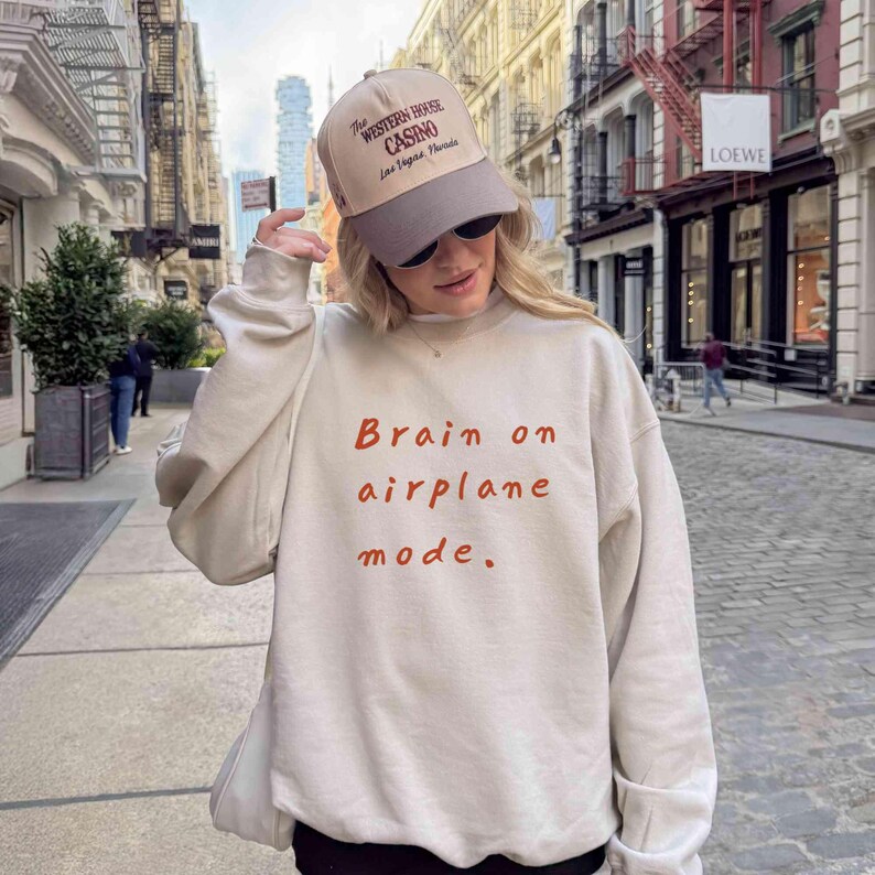 K&ouml;nnte beinhalten: Beiges Sweatshirt mit dem Text "Brain on airplane mode" in roten Buchstaben. Die Person tr&auml;gt eine beige und graue Baseballkappe mit dem Aufdruck "The Western House Casino Las Vegas Florida".