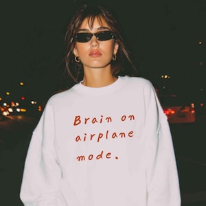 K&ouml;nnte beinhalten: Wei&szlig;es Sweatshirt mit dem roten Schriftzug "Brain on airplane mode". Das Bild wurde nachts aufgenommen, mit verschwommenen Lichtern im Hintergrund. Das Sweatshirt ist ein l&auml;ssiger Stil.