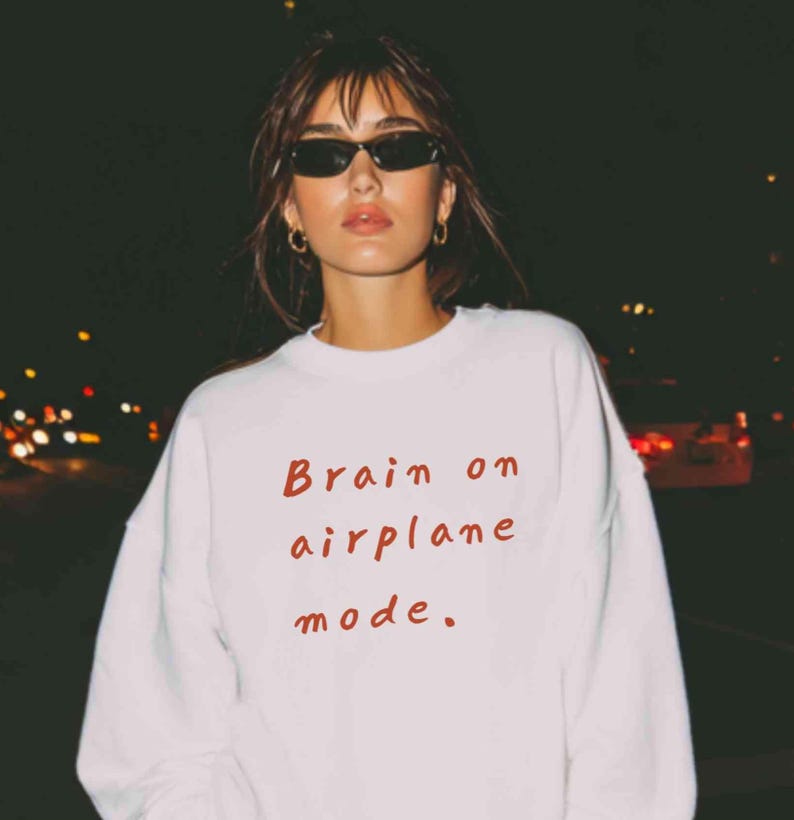 K&ouml;nnte beinhalten: Wei&szlig;es Sweatshirt mit dem roten Schriftzug "Brain on airplane mode". Das Bild wurde nachts aufgenommen, mit verschwommenen Lichtern im Hintergrund. Das Sweatshirt ist ein l&auml;ssiger Stil.