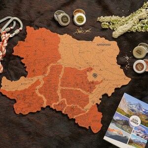 Könnte beinhalten: Eine Korktafelkarte der österreichischen Alpen mit dem Text "ALPENSPINNER" obenauf. Die Karte ist umgeben von kleinen Metallbehältern, Stecknadeln und einem weißen Seil mit Karabinerhaken.