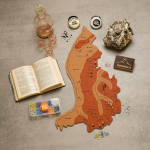 Könnte beinhalten: Eine Korktafelkarte von Österreich mit roten, braunen und weißen Markierungen. Die Karte ist von verschiedenen Gegenständen umgeben, darunter ein Buch, eine Glaskaraffe, kleine Gläser und eine Schachtel Stecknadeln. Die Karte ist mit "Alpenplaner.at" beschriftet.