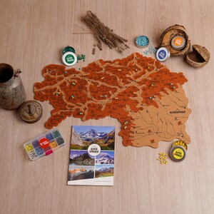 May include: A cork map of a mountainous region with pins marking locations. The map is brown with detailed topographical features. The image includes a brochure, tins of pins, and decorative elements like dried lavender and a metal container.