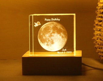 Op de afbeelding: Een heldere glazen kubus met een maan en een ruimteschip erin gegraveerd. De kubus wordt van achteren verlicht, waardoor een gloeiend effect ontstaat. De tekst "Happy Birthday", "Jack" en "March 3, 1999" is ook in de kubus gegraveerd. De kubus staat op een houten voet.