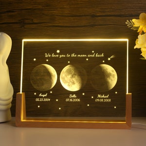 May include: A wooden framed illuminated sign with three crescent moons and the text "We love you to the moon and back". Each moon has a name and date below it: "Angel 05.23.2004", "Bella 07.16.2006", and "Michael 09.08.2008".