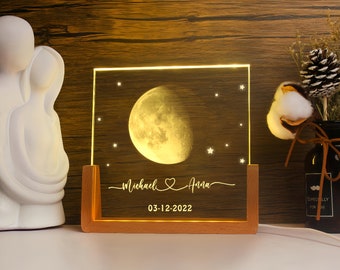 May include: A personalized night light with a moon and stars design. The light has a wooden base and a clear acrylic panel with the names "Michael & Anna" and the date "03-12-2022" engraved. The moon is illuminated with a warm glow.
