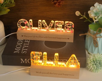 May include: Two illuminated wooden name signs, "OLIVER" and "ELLA", with floral accents. Each name is lit from within, resting on a wooden base with an engraved message. The signs are on a table with decorative elements.