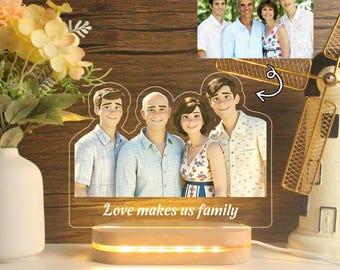 Puede incluir: Una luz nocturna de madera con una silueta de familia de dibujos animados. La silueta está llena de fotos de una familia. La base de la luz nocturna está hecha de madera y tiene un brillo cálido. El texto "Love makes us family" está impreso en la luz nocturna.
