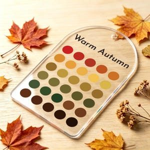 Peut inclure: Une palette de couleurs en acrylique transparent, intitulée "Warm Autumn", avec une série de cercles colorés dans les tons de rouge, orange, jaune, vert et marron. Des feuilles d'automne sont dispersées autour de la palette sur une surface en bois.