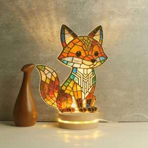 Może przedstawiać: Lampa w kształcie lisa wykonana ze witrażu z drewnianą podstawą. Lis wykonany jest z kolorowych szklanych elementów w odcieniach pomarańczu, żółci, zieleni i niebieskiego. Lampa jest podświetlona, a światło przechodzi przez szkło. Po lewej stronie lampy znajduje się brązowy wazon.