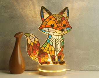 Puede incluir: Una lámpara de zorro de vidrieras con una base de madera. El zorro está hecho de piezas de vidrio de colores en tonos naranja, amarillo, verde y azul. La lámpara está encendida y la luz brilla a través del vidrio. Un jarrón marrón está a la izquierda de la lámpara.