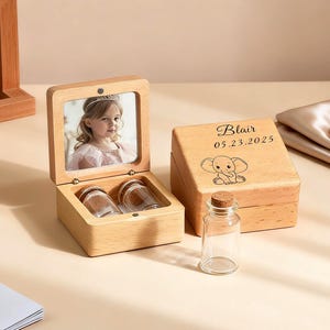 May include: A wooden keepsake box with a photo of a child inside. The box is open, revealing two small glass jars. A second box is closed, with the name "Blair", date, and a baby elephant illustration. A small glass bottle with a cork is in front.