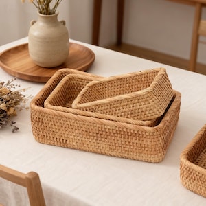 May include: Three woven rectangular baskets in varying sizes are nested together. The baskets are made of a light brown material and have a textured surface. A wooden tray and vase with dried flowers are in the background.