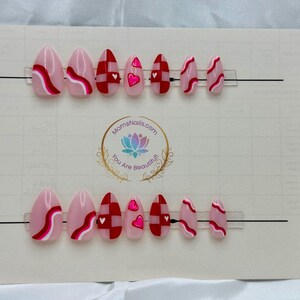 May include: Two sets of almond-shaped press-on nails. Each set features a variety of designs, including pink bases with red and white wavy lines, red and white checkered patterns, and pink nails with heart accents. The nails are displayed on a white card.