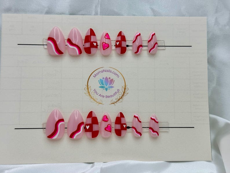 May include: Two sets of almond-shaped press-on nails. Each set features a variety of designs, including pink bases with red and white wavy lines, red and white checkered patterns, and pink nails with heart accents. The nails are displayed on a white card.
