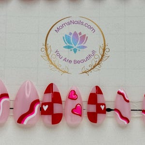 May include: Set of pink and red press-on nails with various designs, including wavy lines, checkered patterns, and heart accents. The nails are oval-shaped and feature a glossy finish. The logo "MomsNails.com You Are Beautiful!" is visible.