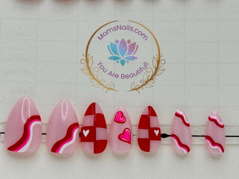 May include: Set of pink and red press-on nails with various designs, including wavy lines, checkered patterns, and heart accents. The nails are oval-shaped and feature a glossy finish. The logo "MomsNails.com You Are Beautiful!" is visible.