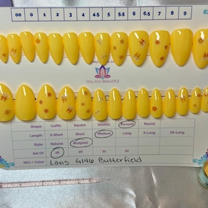 Puede incluir: Un juego de uñas postizas amarillas en forma de almendra con pequeños detalles florales naranjas. Las uñas están dispuestas en una tarjeta blanca con indicadores de tamaño y el texto "You Are Beautiful". El SKU es Lavis G146 Butterfield.
