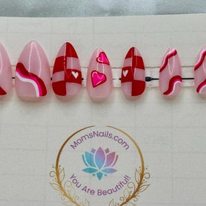 May include: Set of seven almond-shaped press-on nails. The nails feature pink, red, and white designs, including hearts, wavy lines, and checkered patterns. The nails are arranged on a clear plastic strip.