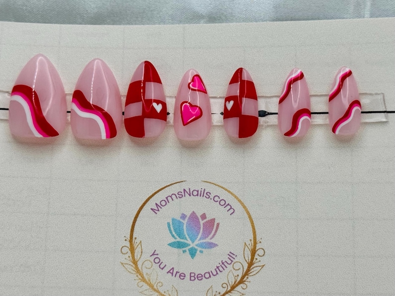 May include: Set of seven almond-shaped press-on nails. The nails feature pink, red, and white designs, including hearts, wavy lines, and checkered patterns. The nails are arranged on a clear plastic strip.