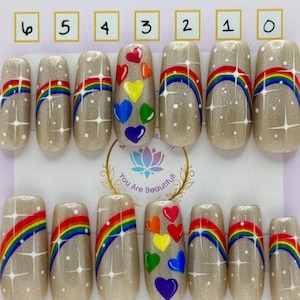 Può includere: Un set di unghie finte con una base beige scintillante. Le unghie presentano arcobaleni dipinti a mano, stelle bianche e cuori colorati. La scheda di presentazione include il testo "Moms Nails" e "You Are Beautiful!" con etichette.