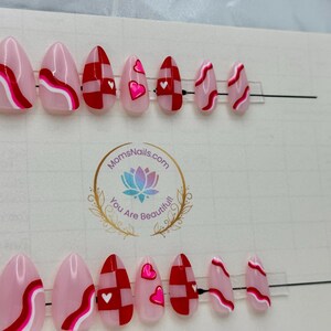 May include: A set of oval-shaped press-on nails with various designs. Some nails feature pink and white wavy lines, while others have red and white checkered patterns or pink hearts. The nails are displayed on a white background.