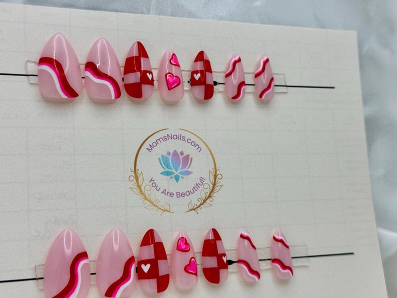 May include: A set of oval-shaped press-on nails with various designs. Some nails feature pink and white wavy lines, while others have red and white checkered patterns or pink hearts. The nails are displayed on a white background.