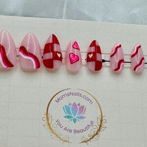 May include: A set of almond-shaped press-on nails with various designs. Some nails feature pink and red abstract patterns, while others have red and white checkered patterns or pink hearts. The nails are arranged on a clear plastic strip.