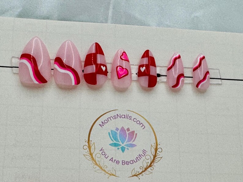 May include: A set of almond-shaped press-on nails with various designs. Some nails feature pink and red abstract patterns, while others have red and white checkered patterns or pink hearts. The nails are arranged on a clear plastic strip.