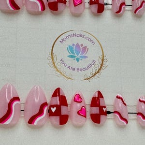 May include: Oval-shaped press-on nails in a variety of designs. Some nails feature a pink base with red and white wavy lines. Others have a red and white checkerboard pattern with small white hearts. The nails are arranged on a clear plastic holder.