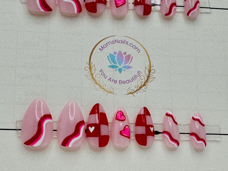 May include: Oval-shaped press-on nails in a variety of designs. Some nails feature a pink base with red and white wavy lines. Others have a red and white checkerboard pattern with small white hearts. The nails are arranged on a clear plastic holder.