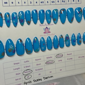 Puede incluir: Un conjunto de uñas postizas azules en forma de almendra y de longitud media, con diseños florales. Las uñas están dispuestas sobre una tarjeta blanca con indicadores de tamaño y el texto "MomsNails.com" y "You Are Beautiful". El conjunto está etiquetado como "Après Dusky Denim".