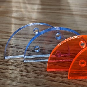 Könnte beinhalten: Drei halbkreisförmige, transparente Objekte mit Löchern. Zwei sind blau und eines ist orange. Sie sind in einer Reihe auf einer Holzoberfläche angeordnet. Die Objekte scheinen aus Acryl oder einem ähnlichen Material zu bestehen.