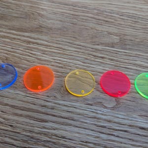 Könnte beinhalten: Fünf transparente, farbige Acrylkreise in Blau, Orange, Gelb, Rot und Grün. Jede Scheibe hat zwei kleine Löcher. Die Objekte sind diagonal auf einer Holzoberfläche angeordnet.