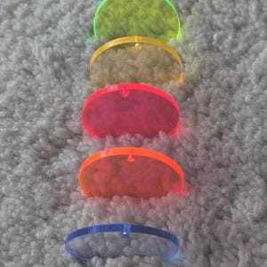 Könnte beinhalten: Fünf farbige, transluzente, halbkreisförmige Acrylscheiben in vertikaler Anordnung. Die Farben sind von oben nach unten: Neongrün, Gelb, Pink, Orange und Blau. Jede Scheibe hat oben ein kleines Loch. Auf einer strukturierten, hellen Oberfläche.